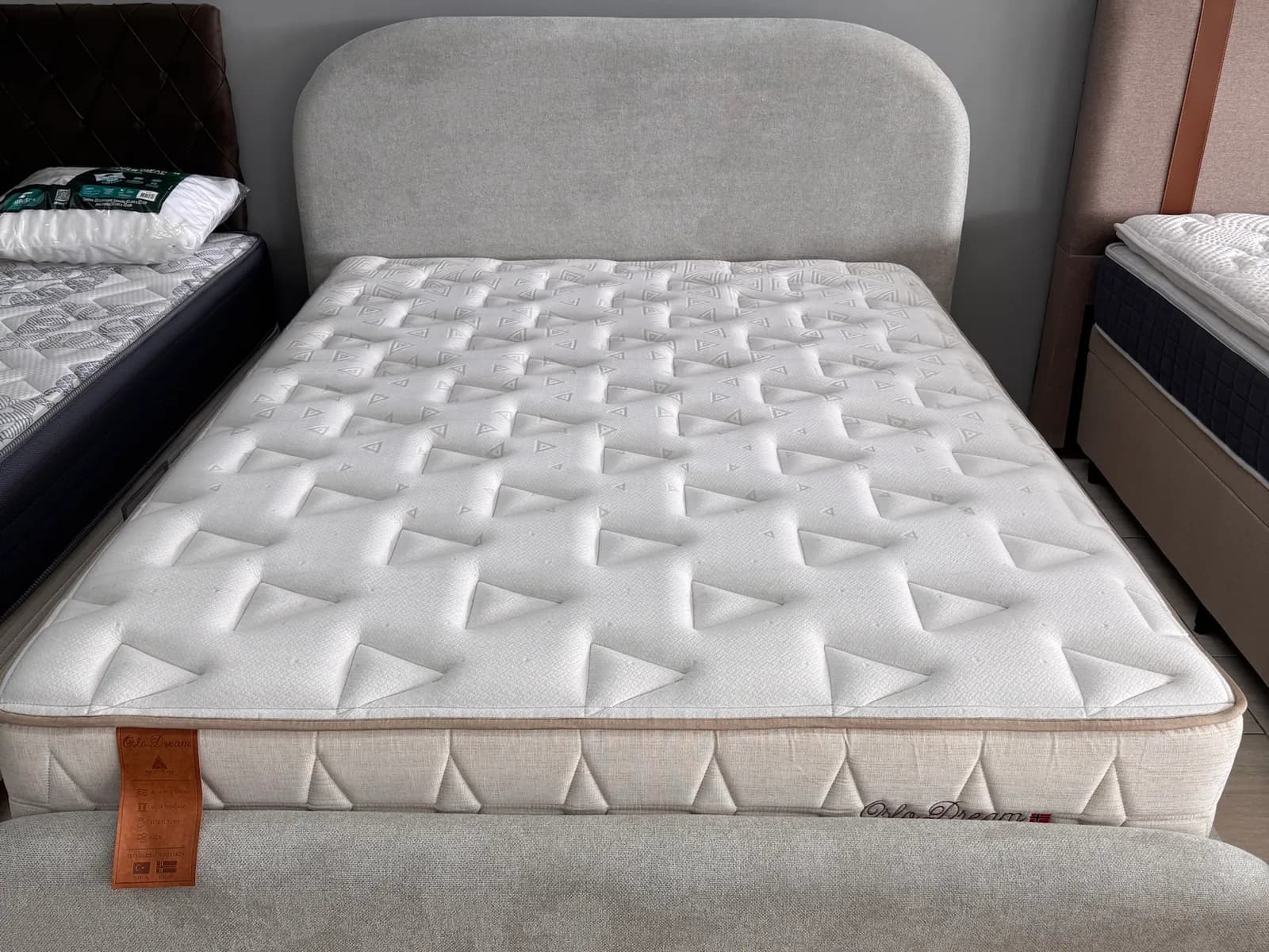 Colchão Oslo Dream em cama estofada cinza com cabeceira arredondada no showroom da Decorelar Colchões, em Inoã, Maricá
